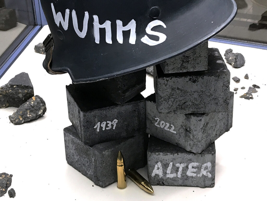 Ansicht Nah - Helm mit Wumms Jahreszahlen Kriegsbeginn, Patronen, Alter auf Stein geschrieben - Kunstinstallation - Krisen-Narrative - präsentiert von Warte Positionen, U-Bahnsteig Bockenheimer Warte