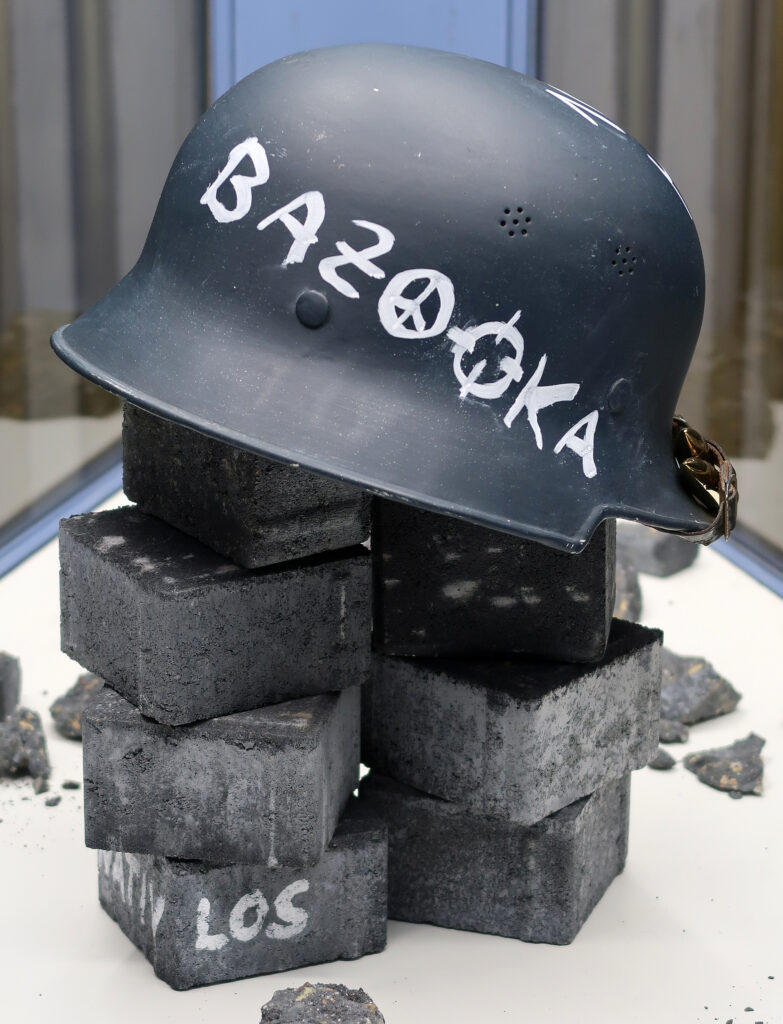 Ansicht Nah - Helm mit Aufschrift Bazooka, auf Steinen platziert, Los auf Stein geschrieben - Kunstinstallation - Krisen-Narrative - präsentiert von Warte Positionen, U-Bahnsteig Bockenheimer Wart
