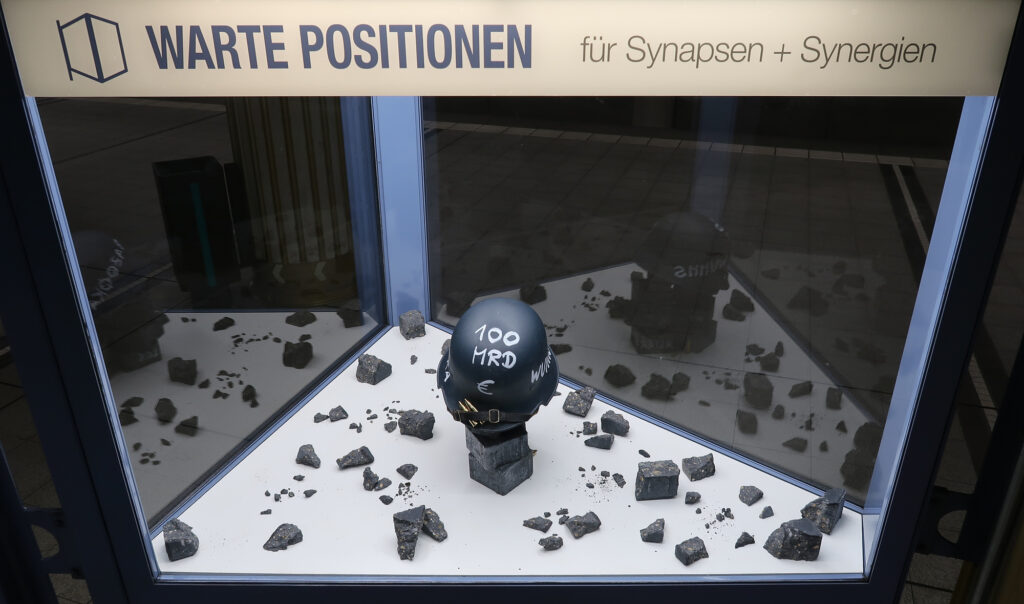 Ansicht Totale - Helm mit Aufschrift Bazooka, auf Steinen platziert, Los auf Stein geschrieben - Kunstinstallation - Krisen-Narrative - präsentiert von Warte Positionen, U-Bahnsteig Bockenheimer Wart